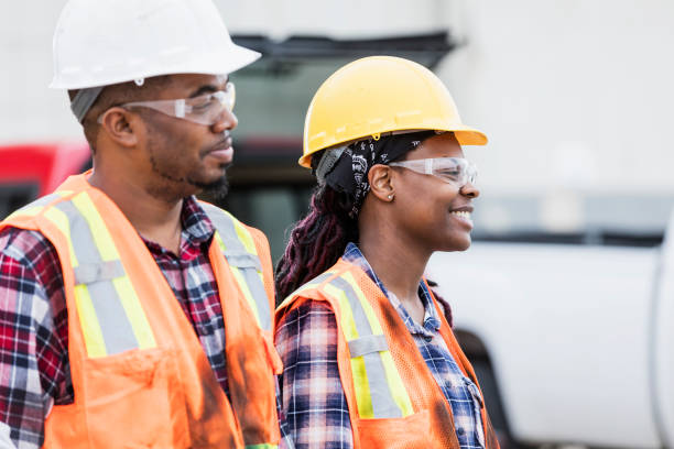 Two construction workers standing outdoors, wearing hard hats, safety glasses, reflective vest and plaid shirts. The focus is on the young African-American woman. Her coworker is in his 30s.