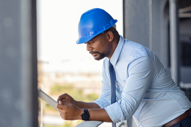 Mature manager using digital tablet on construction site. Serious african engineer checking email at building site wearing blue hardhat. Architect working on digital tablet leaning at balustrade.